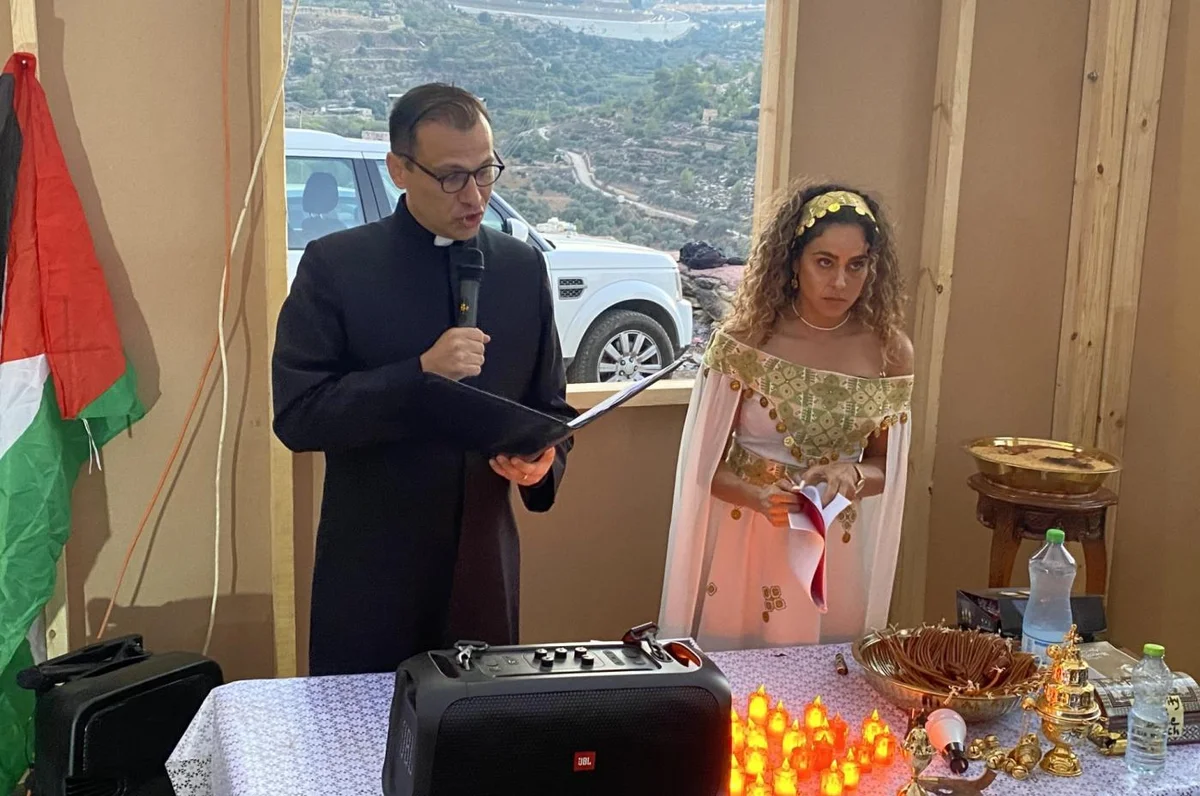A priest leads a prayer service beside Alice Kisiya in traditional embroidered dress, overlooking the Al-Makhrour valley